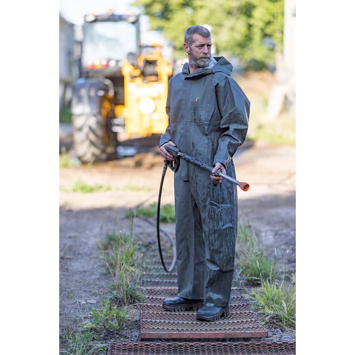 Man in a waterproof full body suit holding a hose on a construction site and farm with machinery in the background